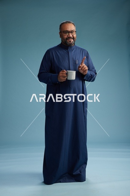 بورتريه لرجل عربي سعودي  خليجي بثوب أزرق يحمل كوب قهوة ويبتسم, الاستمتاع بالمشروب الساخن, الجسم بالطول الكامل, خلفيه زرقاء