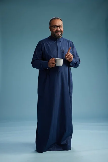 بورتريه لرجل عربي سعودي  خليجي بثوب أزرق يحمل كوب قهوة ويبتسم, الاستمتاع بالمشروب الساخن, الجسم بالطول الكامل, خلفيه زرقاء
