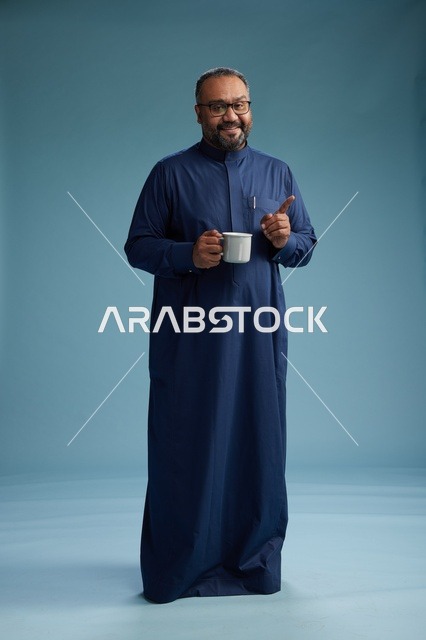 بورتريه لرجل عربي سعودي  خليجي بثوب أزرق يحمل كوب قهوة ويبتسم, الاستمتاع بالمشروب الساخن, الجسم بالطول الكامل, خلفيه زرقاء