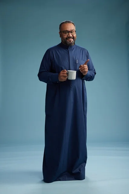 A portrait of a Saudi Arabian Gulf man in a blue thobe holding a cup of coffee and smiling, enjoying the hot drink, full-body shot, with a blue background.