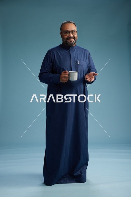 بورتريه لرجل عربي سعودي  خليجي بثوب أزرق يحمل كوب قهوة ويبتسم, الاستمتاع بالمشروب الساخن, الجسم بالطول الكامل, خلفيه زرقاء