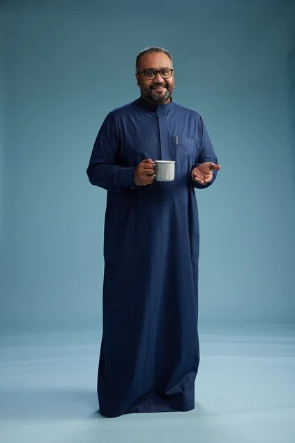 A portrait of a Saudi Arabian Gulf man in a blue thobe holding a cup of coffee and smiling, enjoying the hot drink, full-body shot, with a blue background.