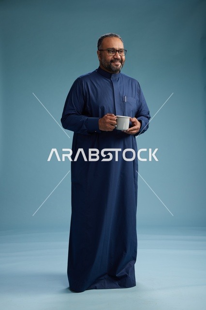 بورتريه لرجل عربي سعودي  خليجي بثوب أزرق يحمل كوب قهوة ويبتسم, الاستمتاع بالمشروب الساخن, الجسم بالطول الكامل, خلفيه زرقاء
