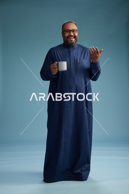 بورتريه لرجل عربي سعودي  خليجي بثوب أزرق يحمل كوب قهوة ويبتسم, الاستمتاع بالمشروب الساخن, الجسم بالطول الكامل, خلفيه زرقاء