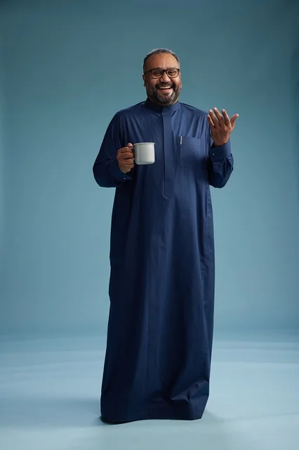 بورتريه لرجل عربي سعودي  خليجي بثوب أزرق يحمل كوب قهوة ويبتسم, الاستمتاع بالمشروب الساخن, الجسم بالطول الكامل, خلفيه زرقاء