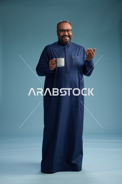 بورتريه لرجل عربي سعودي  خليجي بثوب أزرق يحمل كوب قهوة ويبتسم, الاستمتاع بالمشروب الساخن, الجسم بالطول الكامل, خلفيه زرقاء