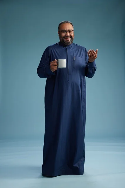 A portrait of a Saudi Arabian Gulf man in a blue thobe holding a cup of coffee and smiling, enjoying the hot drink, full-body shot, with a blue background.