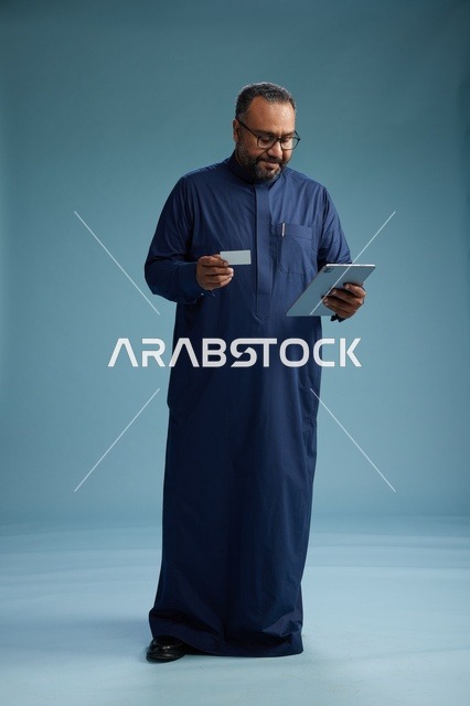 بورتريه لرجل عربي سعودي  خليجي يرتدي ثوباً أزرق يقرأ بطاقة عمل مستخدماً جهازاً لوحياً, استخدام البطاقه البنكيه, كامل الجسم, خلفيه زرقاء