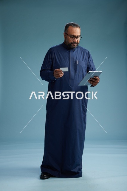 بورتريه لرجل عربي سعودي  خليجي يرتدي ثوباً أزرق يقرأ بطاقة عمل مستخدماً جهازاً لوحياً, استخدام البطاقه البنكيه, كامل الجسم, خلفيه زرقاء