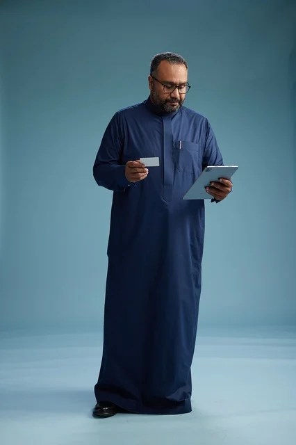 A portrait of a Saudi Arabian Gulf man wearing a blue thobe reading a business card while using a tablet, using a bank card, full body, blue background.