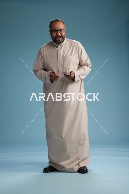 بورتريه لرجل  عربي سعودي خليجي يرتدي ثوب بيج ويحمل هاتفاً ذكياً, استخدام جهاز الجوال, كامل الجسم, خلفيه زرقاء