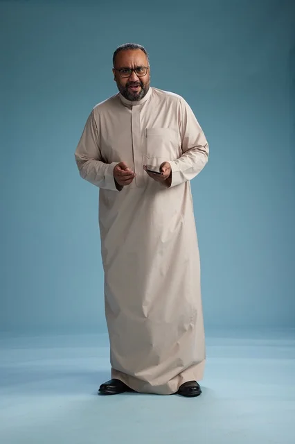 بورتريه لرجل  عربي سعودي خليجي يرتدي ثوب بيج ويحمل هاتفاً ذكياً, استخدام جهاز الجوال, كامل الجسم, خلفيه زرقاء
