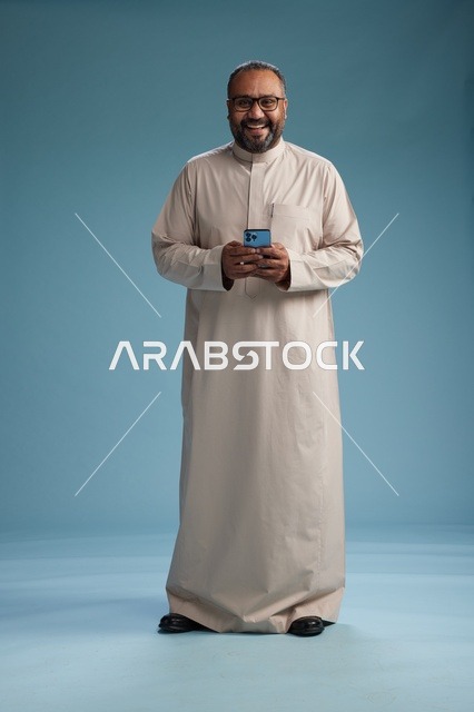بورتريه لرجل  عربي سعودي خليجي يرتدي ثوب بيج ويحمل هاتفاً ذكياً, استخدام جهاز الجوال, كامل الجسم, خلفيه زرقاء