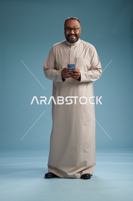 A portrait of an Arab Saudi Gulf man wearing a beige thobe and holding a smartphone, using a mobile device, full body, blue background.