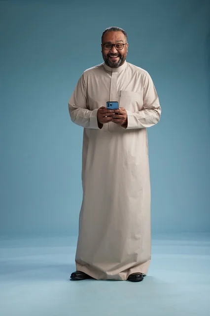 A portrait of an Arab Saudi Gulf man wearing a beige thobe and holding a smartphone, using a mobile device, full body, blue background.