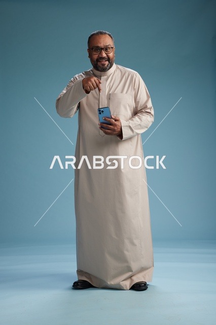 بورتريه لرجل  عربي سعودي خليجي يرتدي ثوب بيج ويحمل هاتفاً ذكياً, استخدام جهاز الجوال, كامل الجسم, خلفيه زرقاء