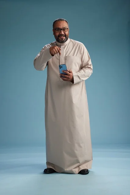 A portrait of an Arab Saudi Gulf man wearing a beige thobe and holding a smartphone, using a mobile device, full body, blue background.
