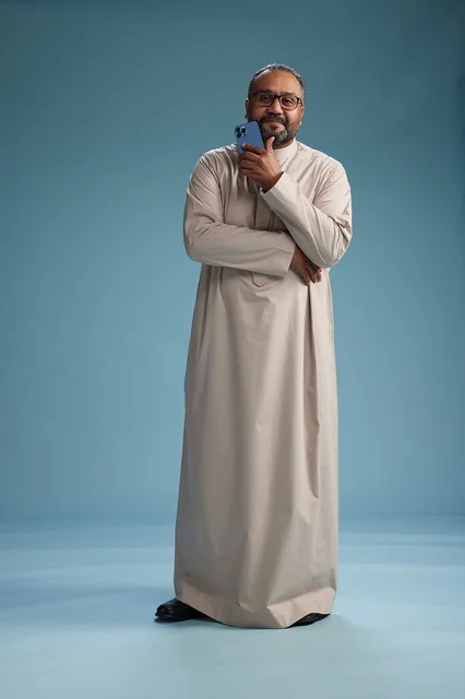 A portrait of an Arab Saudi Gulf man wearing a beige thobe and holding a smartphone, using a mobile device, full body, blue background.
