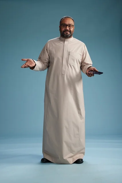 A portrait of an Arab Saudi Gulf man wearing a beige thobe and holding a smartphone, using a mobile device, full body, blue background.