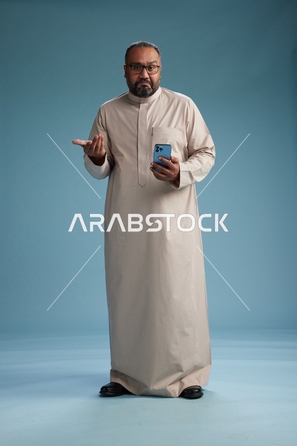بورتريه لرجل  عربي سعودي خليجي يرتدي ثوب بيج ويحمل هاتفاً ذكياً, استخدام جهاز الجوال, كامل الجسم, خلفيه زرقاء