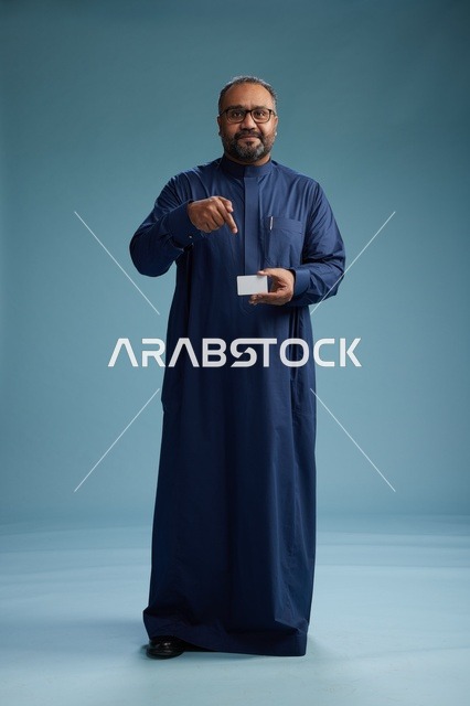 بورتريه لرجل عربي  سعودي  خليجي يرتدي ثوباً أزرق يمسك بيده بطاقة بيضاء فارغه, الجسم بالكامل, خلفيه زرقاء