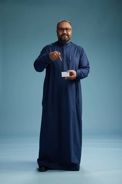 A portrait of an Arab man, Saudi, Gulf, wearing a blue thobe, holding a blank white card in his hand, full body, blue background.