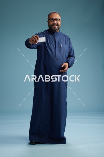 بورتريه لرجل عربي  سعودي  خليجي يرتدي ثوباً أزرق يمسك بيده بطاقة بيضاء فارغه, الجسم بالكامل, خلفيه زرقاء