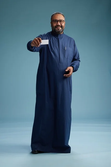 بورتريه لرجل عربي  سعودي  خليجي يرتدي ثوباً أزرق يمسك بيده بطاقة بيضاء فارغه, الجسم بالكامل, خلفيه زرقاء