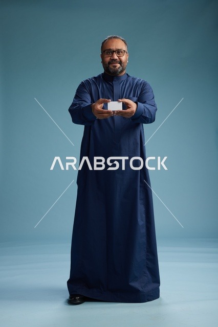 بورتريه لرجل عربي  سعودي  خليجي يرتدي ثوباً أزرق يمسك بيده بطاقة بيضاء فارغه, الجسم بالكامل, خلفيه زرقاء