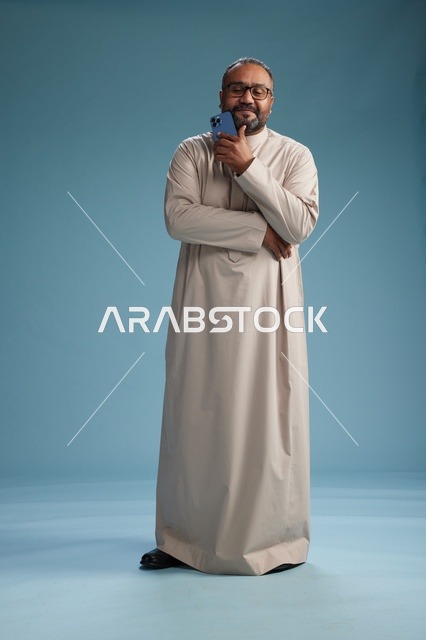 بورتريه لرجل  عربي سعودي خليجي يرتدي ثوب بيج ويحمل هاتفاً ذكياً, استخدام جهاز الجوال, كامل الجسم, خلفيه زرقاء