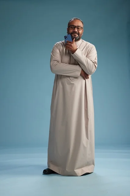 A portrait of an Arab Saudi Gulf man wearing a beige thobe and holding a smartphone, using a mobile device, full body, blue background.