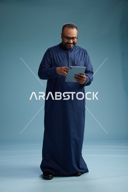 بورتريه لرجل عربي سعودي  خليجي بثوب أزرق يستخدم جهاز لوحي, استخدام التابليت المحمول, العمل اونلاين, الجسم بالكامل, خلفيه زرقاء