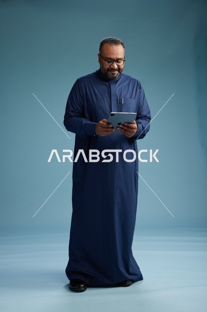 Portrait of a Saudi Arabian Gulf man in a blue thobe using a tablet, using a mobile tablet, working online, full body, blue background.