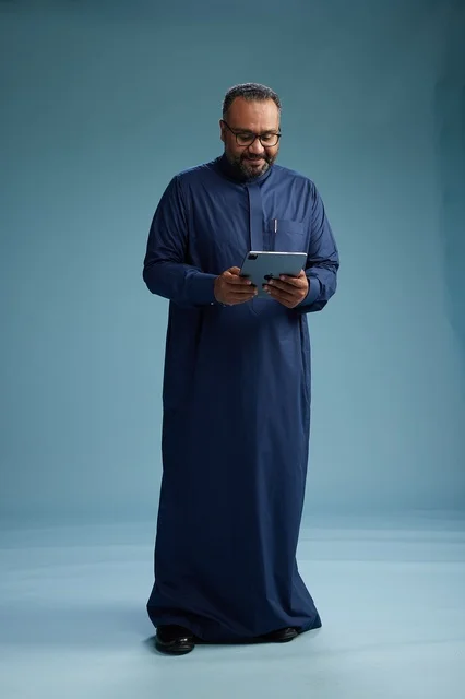 بورتريه لرجل عربي سعودي  خليجي بثوب أزرق يستخدم جهاز لوحي, استخدام التابليت المحمول, العمل اونلاين, الجسم بالكامل, خلفيه زرقاء