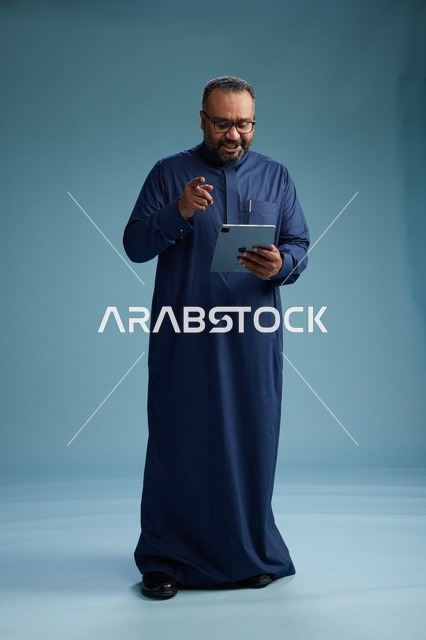 بورتريه لرجل عربي سعودي  خليجي بثوب أزرق يستخدم جهاز لوحي, استخدام التابليت المحمول, العمل اونلاين, الجسم بالكامل, خلفيه زرقاء
