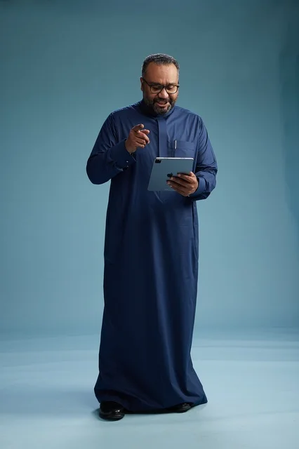 بورتريه لرجل عربي سعودي  خليجي بثوب أزرق يستخدم جهاز لوحي, استخدام التابليت المحمول, العمل اونلاين, الجسم بالكامل, خلفيه زرقاء
