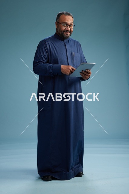 بورتريه لرجل عربي سعودي  خليجي بثوب أزرق يستخدم جهاز لوحي, استخدام التابليت المحمول, العمل اونلاين, الجسم بالكامل, خلفيه زرقاء