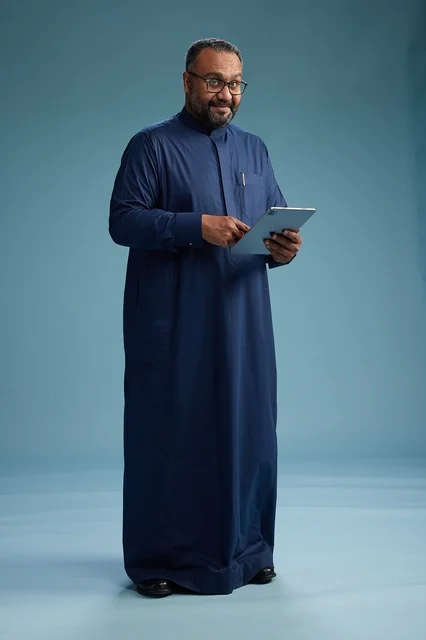 Portrait of a Saudi Arabian Gulf man in a blue thobe using a tablet, using a mobile tablet, working online, full body, blue background.