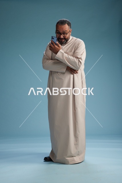 بورتريه لرجل  عربي سعودي خليجي يرتدي ثوب بيج ويحمل هاتفاً ذكياً, استخدام جهاز الجوال, كامل الجسم, خلفيه زرقاء