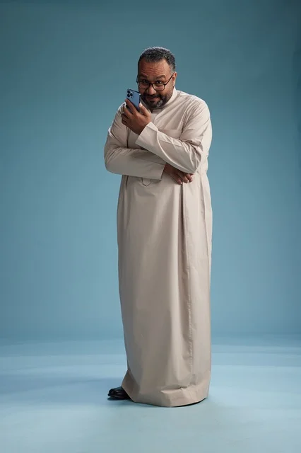 A portrait of an Arab Saudi Gulf man wearing a beige thobe and holding a smartphone, using a mobile device, full body, blue background.