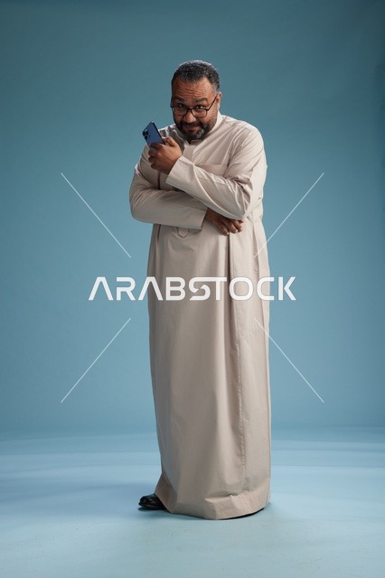 بورتريه لرجل  عربي سعودي خليجي يرتدي ثوب بيج ويحمل هاتفاً ذكياً, استخدام جهاز الجوال, كامل الجسم, خلفيه زرقاء
