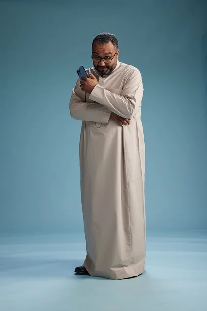 A portrait of an Arab Saudi Gulf man wearing a beige thobe and holding a smartphone, using a mobile device, full body, blue background.