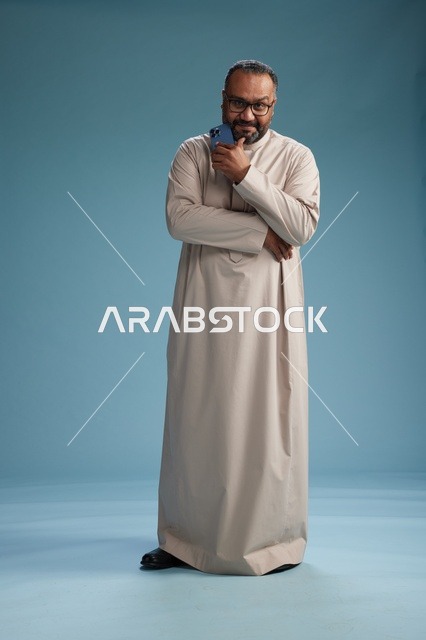 بورتريه لرجل  عربي سعودي خليجي يرتدي ثوب بيج ويحمل هاتفاً ذكياً, استخدام جهاز الجوال, كامل الجسم, خلفيه زرقاء
