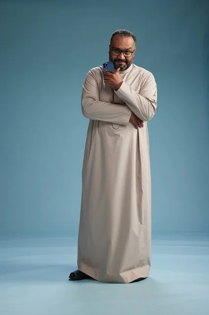 A portrait of an Arab Saudi Gulf man wearing a beige thobe and holding a smartphone, using a mobile device, full body, blue background.