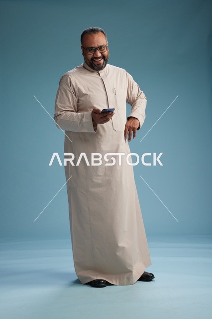 بورتريه لرجل  عربي سعودي خليجي يرتدي ثوب بيج ويحمل هاتفاً ذكياً, استخدام جهاز الجوال, كامل الجسم, خلفيه زرقاء