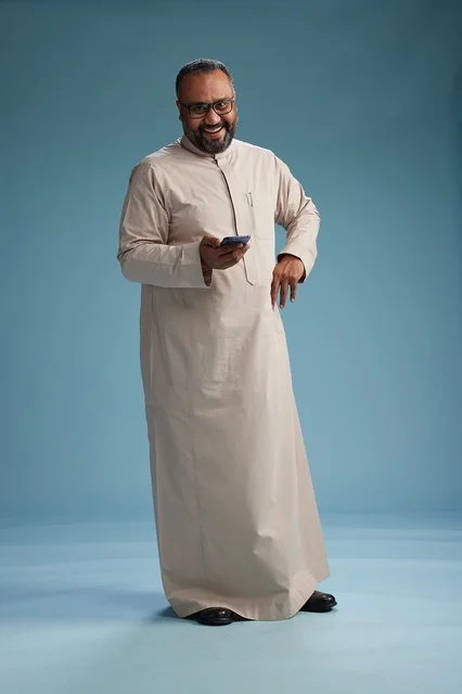 بورتريه لرجل  عربي سعودي خليجي يرتدي ثوب بيج ويحمل هاتفاً ذكياً, استخدام جهاز الجوال, كامل الجسم, خلفيه زرقاء