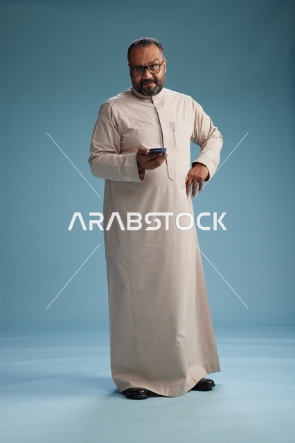 بورتريه لرجل  عربي سعودي خليجي يرتدي ثوب بيج ويحمل هاتفاً ذكياً, استخدام جهاز الجوال, كامل الجسم, خلفيه زرقاء