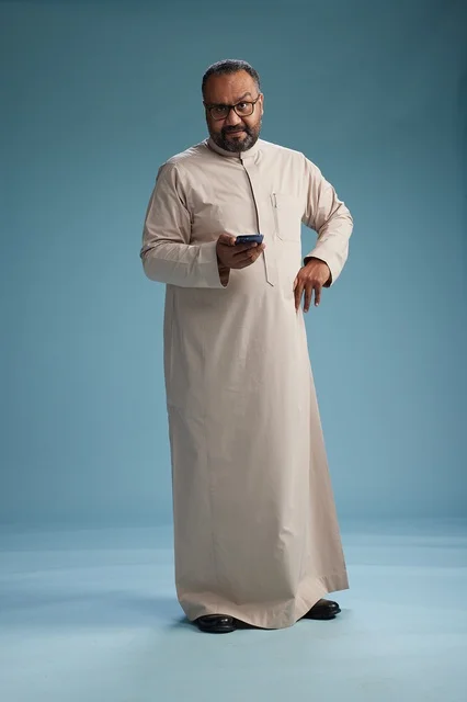 A portrait of an Arab Saudi Gulf man wearing a beige thobe and holding a smartphone, using a mobile device, full body, blue background.