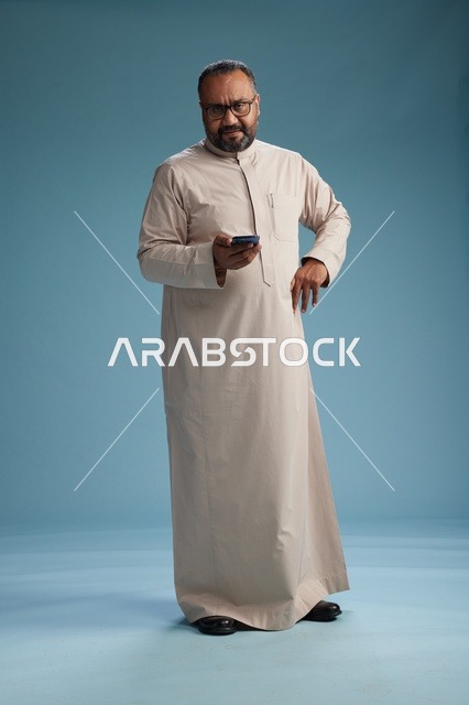بورتريه لرجل  عربي سعودي خليجي يرتدي ثوب بيج ويحمل هاتفاً ذكياً, استخدام جهاز الجوال, كامل الجسم, خلفيه زرقاء