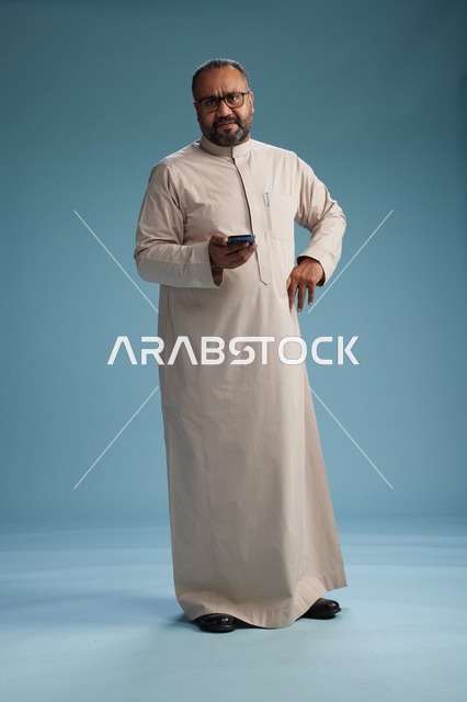 A portrait of an Arab Saudi Gulf man wearing a beige thobe and holding a smartphone, using a mobile device, full body, blue background.